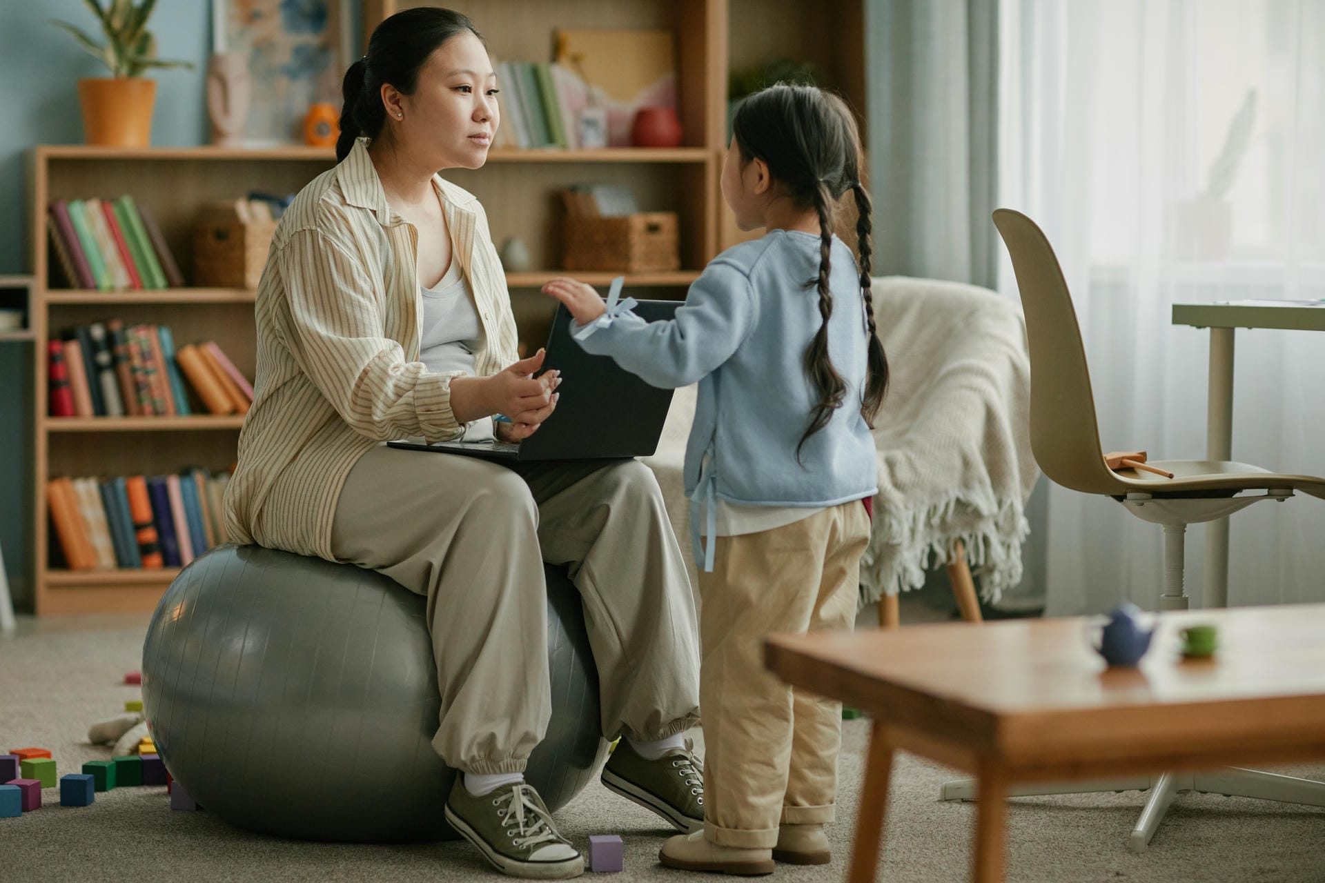 Asian pregnant woman sitting on exercise ball using laptop while spending time with young Asian child standing nearby in living room, engaging in leisure activity together at home