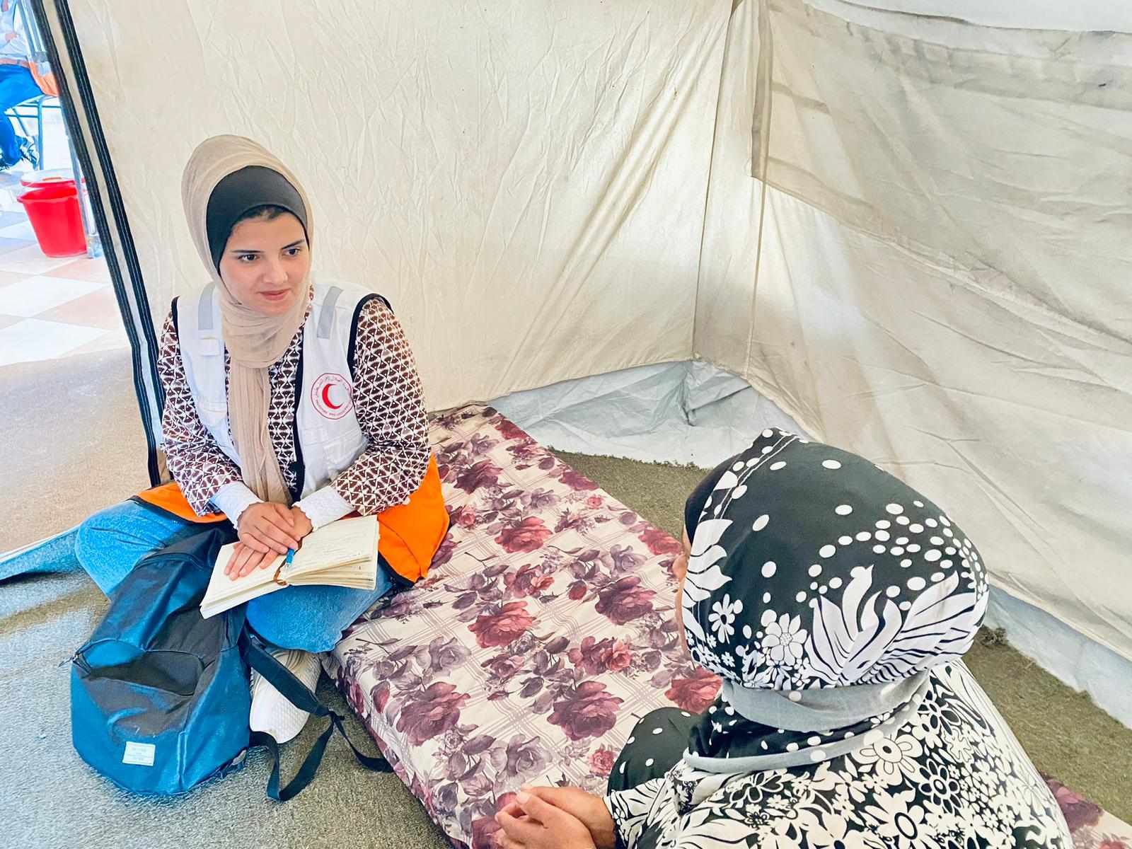 Red Crescent volunteer speaking with a woman inside a relief tent while taking notes during a humanitarian visit